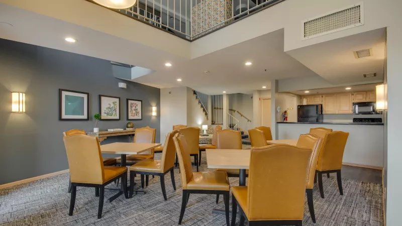 Indoor common space with several round tables, light brown chairs, a small kitchen, and a loft balcony.