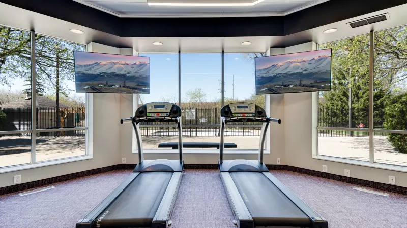 Two treadmills in a gym face large windows with an outdoor view; TVs above display vibrant mountain landscapes.