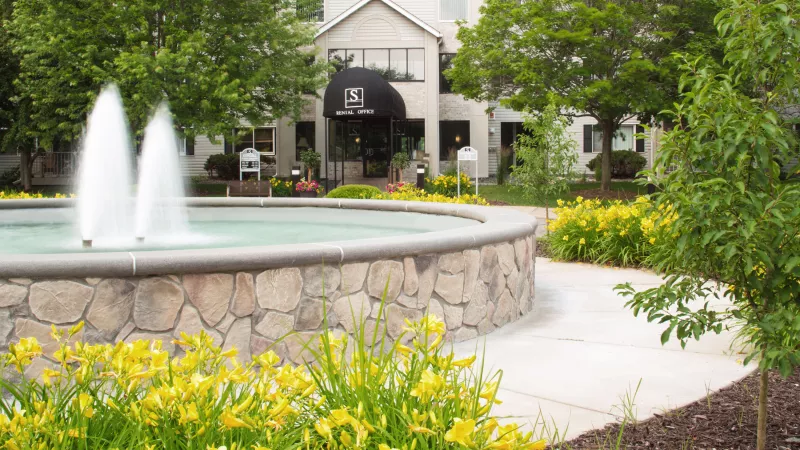 Stone fountain with water streams in front of a building entrance, surrounded by green trees and yellow flowers.