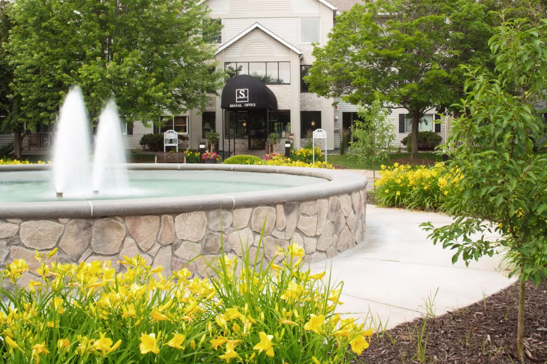 Stone fountain with water streams in front of a building entrance, surrounded by green trees and yellow flowers.