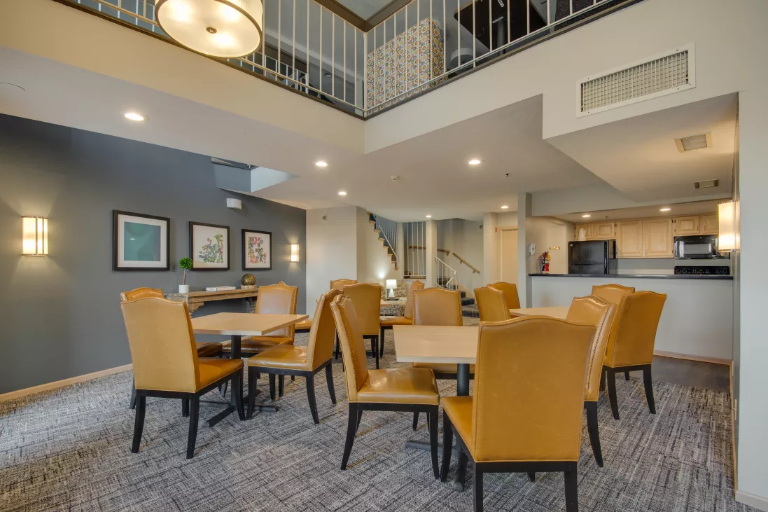 Common lounge with multiple tables, tan chairs, a high ceiling, and an upper balcony. A kitchenette is visible.